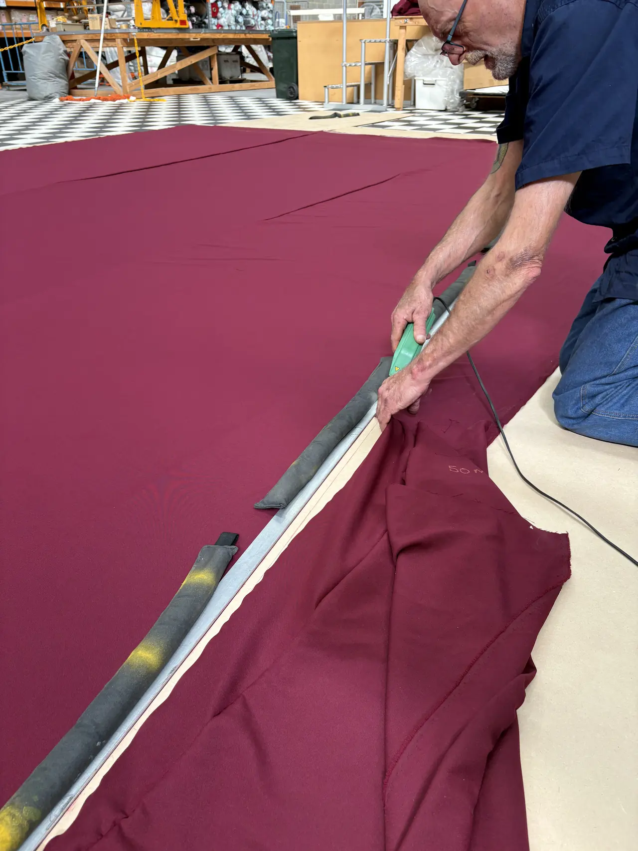 A TSA drape maker kneeling on burgundy cloth, cutting along a metal straight edge with a hot knife